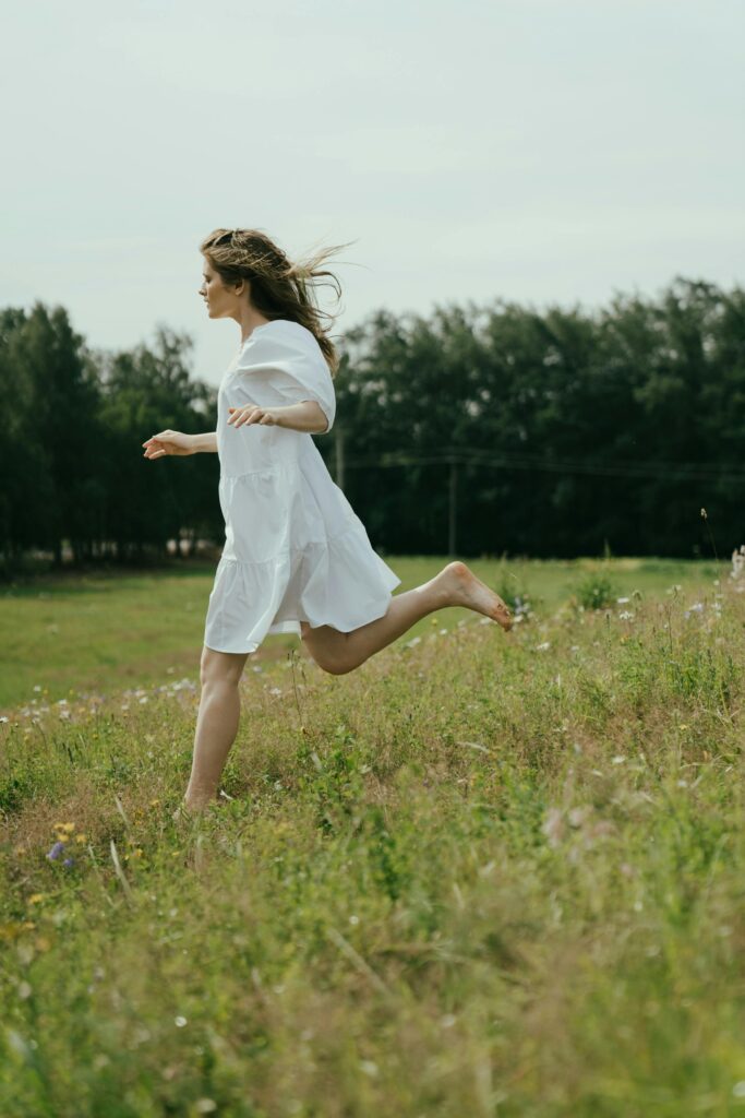 pexels-photo-5615908-5615908 Caucasian woman in a white dress running barefoot in a grassy countryside field.